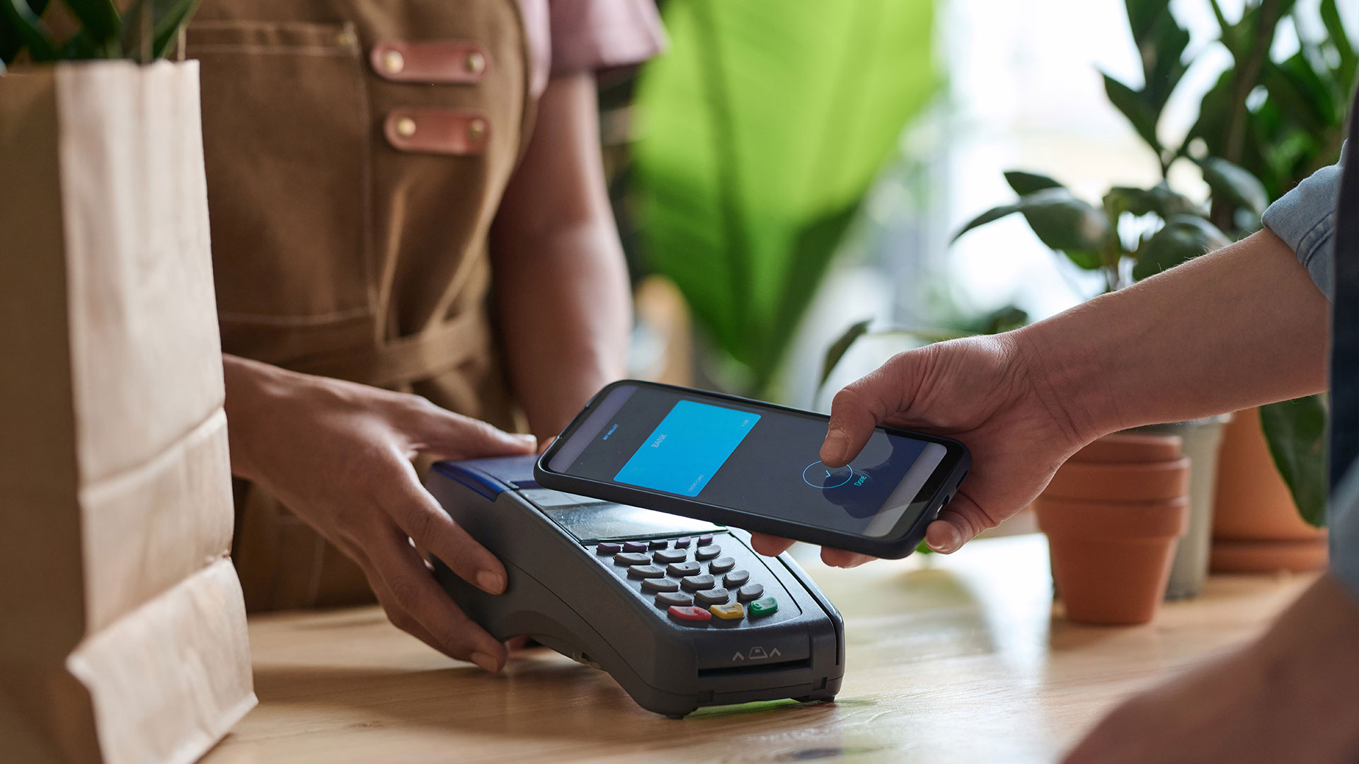 Cliente paga con smartphone contactless in garden center tra piante verdi - il mercato del giardinaggio in Italia vale 3,25 miliardi di euro